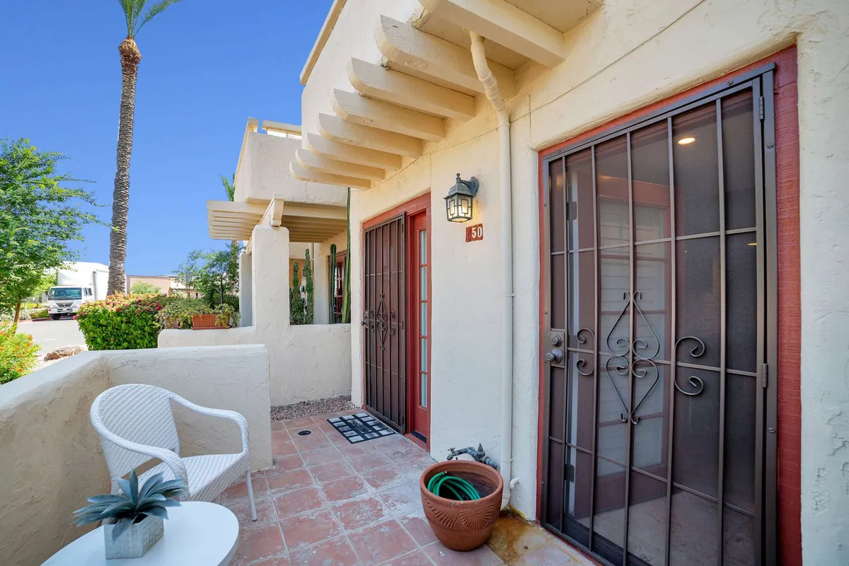 The inviting exterior features decorative double doors, a white wicker chair, and a shaded brick pathway. This cozy space suggests relaxing moments outdoors and an easy transition from home to exploring neighborhood attractions.