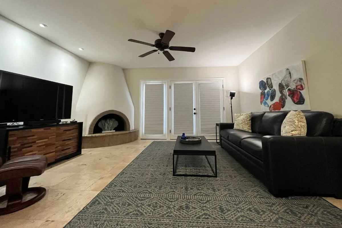 A modern living room featuring a black leather sofa, a large television above a sleek fireplace, and prominent sliding glass doors that lead out to an outdoor space. Artwork on the wall adds personality, and the space is well-lit and cozy, perfect for relaxing or entertaining.