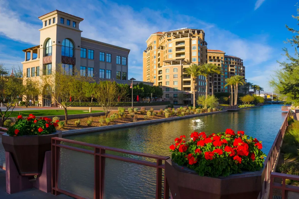 A striking view of waterfront landscaping with red flowers, modern architectural elements, and swaying palm trees. The setting is lively and open, hinting at various outdoor activities and communal enjoyment.