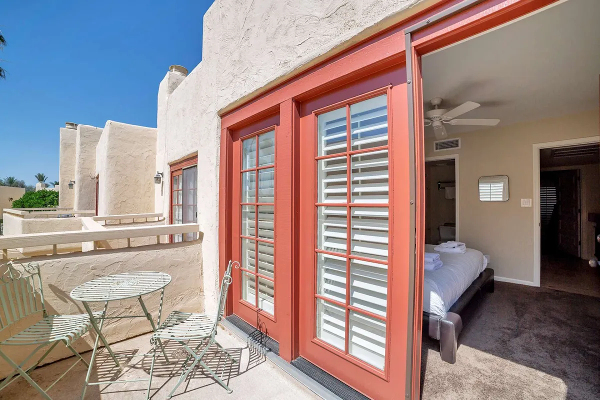An exterior shot of the front patio, featuring a relaxing outdoor seating area with red-framed doors and a partial view into the interior. The scene is bright and serene, hinting at mountain views and a seamless indoor-outdoor connection.