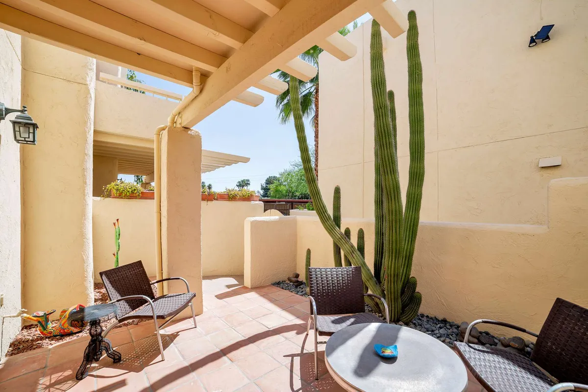 A private patio featuring a simple seating area and table, framed by desert landscaping and a cactus. The setting invites morning coffee, reading, or a casual meal in serene, sunlit surroundings.