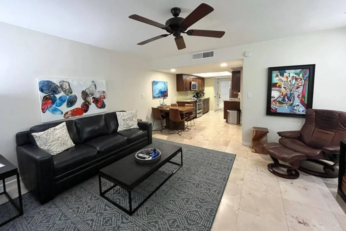 The image showcases a stylish, open-plan living area with a sleek black leather sofa, vibrant modern artwork, and a bright, inviting dining space. A plush recliner chair sits beside a contemporary coffee table, creating the perfect blend of comfort and sophistication. The overall space feels fresh, welcoming, and ideal for both relaxing and entertaining.