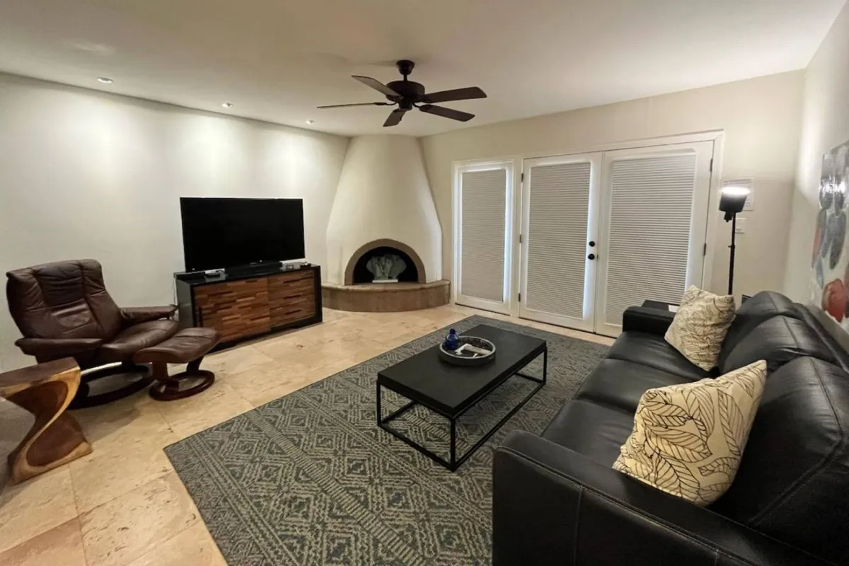 A stylish living room scene with a plush black sofa, a large flat-screen TV positioned above an elegant fireplace, and a unique armchair. The intimate setting invites both group conversation and quiet relaxation by the fire.